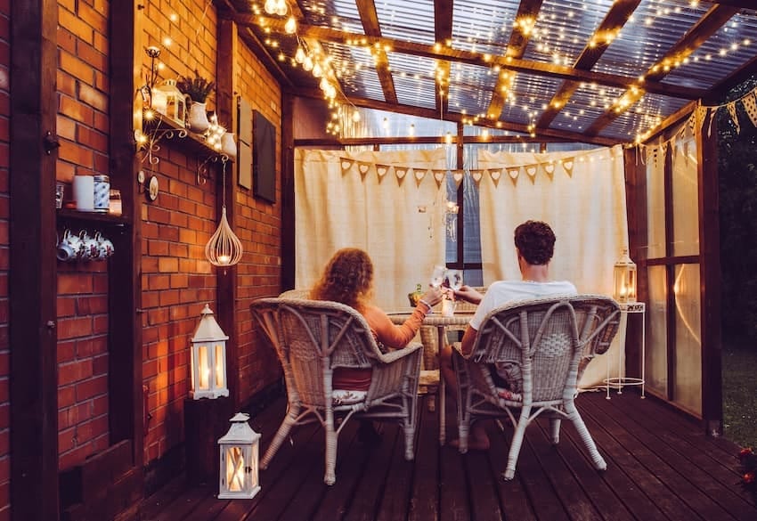 Outdoor-Living im eigenen Garten:  Paar sitzt am Abend neben Hauswand auf einer überdachten Holzterrasse am Tisch beleuchtet von Kerzen und Lichterketten