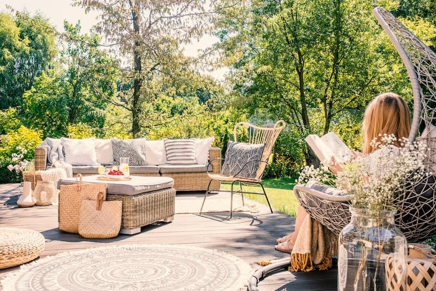 outdoor-living im eigenen Garten: Bild zeigt Terrassendeck auf dem sich Korbmöbel und ein hängender Korbsessel worin lesende Frau im Sonnenschein sitzt