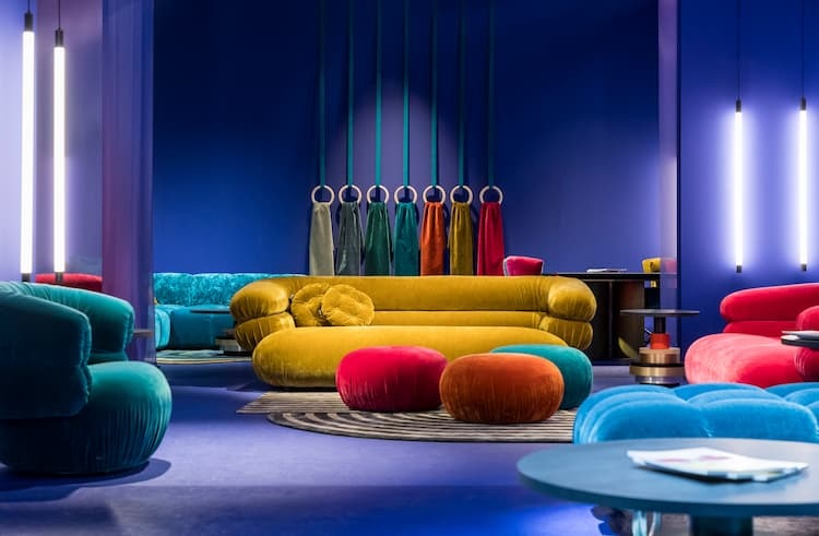 5 Wohntrends rund um Deine Wohnung Sofa von Bretz Bild zeigt Sofas in Gelb, rot und türkis, davor runde Sitzpolsterhocker in rot, türkis braun auf violettem Teppich