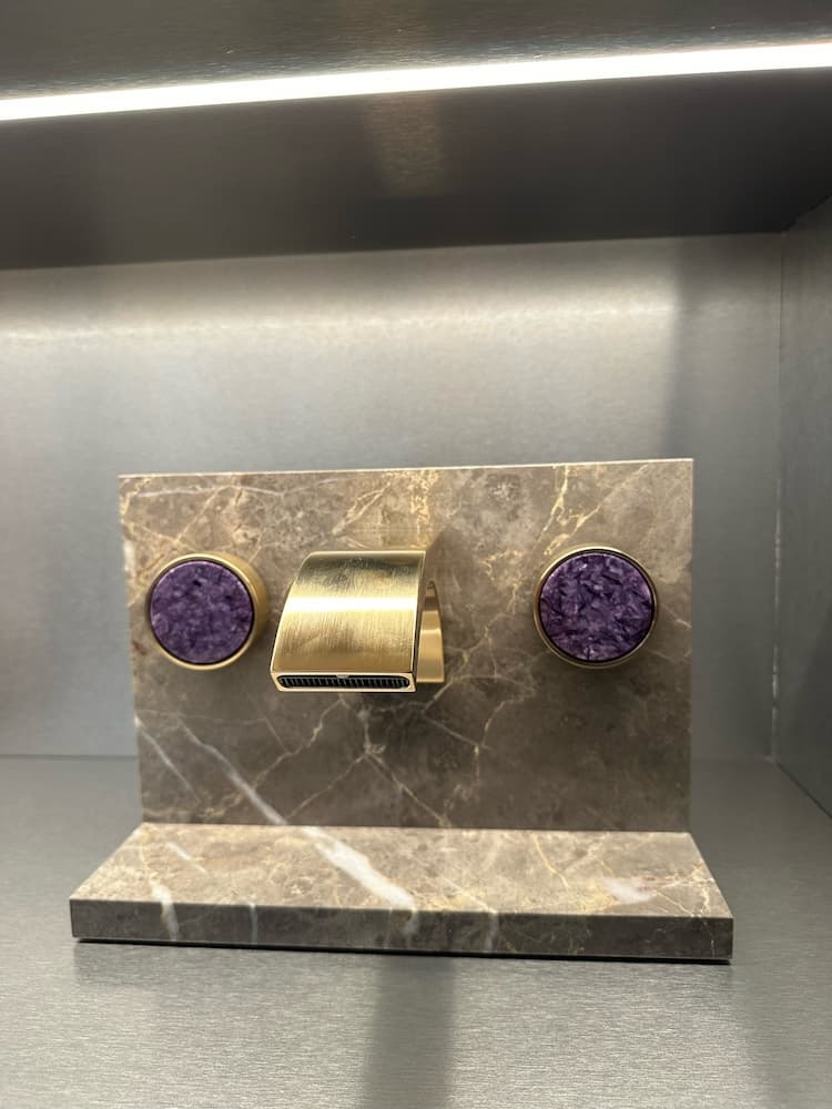 Bild zeigt Gold glänzende geschwungenen breiten Wasserhahn mit zwei runden in violetter Marmoroptik gehaltenen Armaturen an Marmorwand von Dornbracht