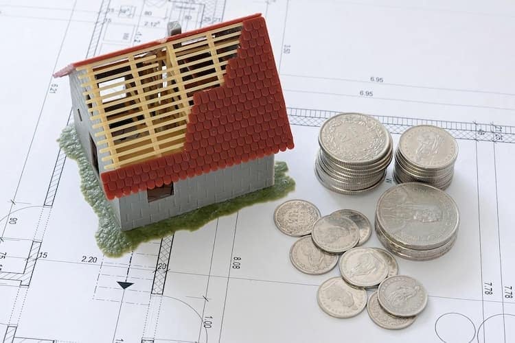 der Hausbau und das liebe Geld: kleines Legohaus steht auf Planskizze, daneben stehen Stapel mit Münzen.