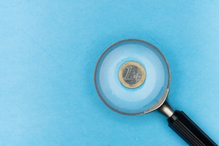 Bild zeigt Euromünze, die auf blauem Untergrund liegt und auf der eine Lupe liegt.