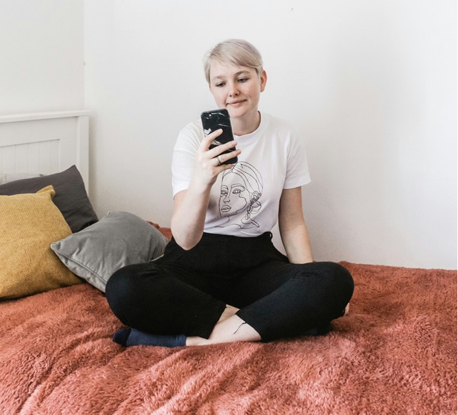 Bild zeigt junge Frau, die zufrieden auf einem Bettsofa sitzt und in ihr handy schreibt. 